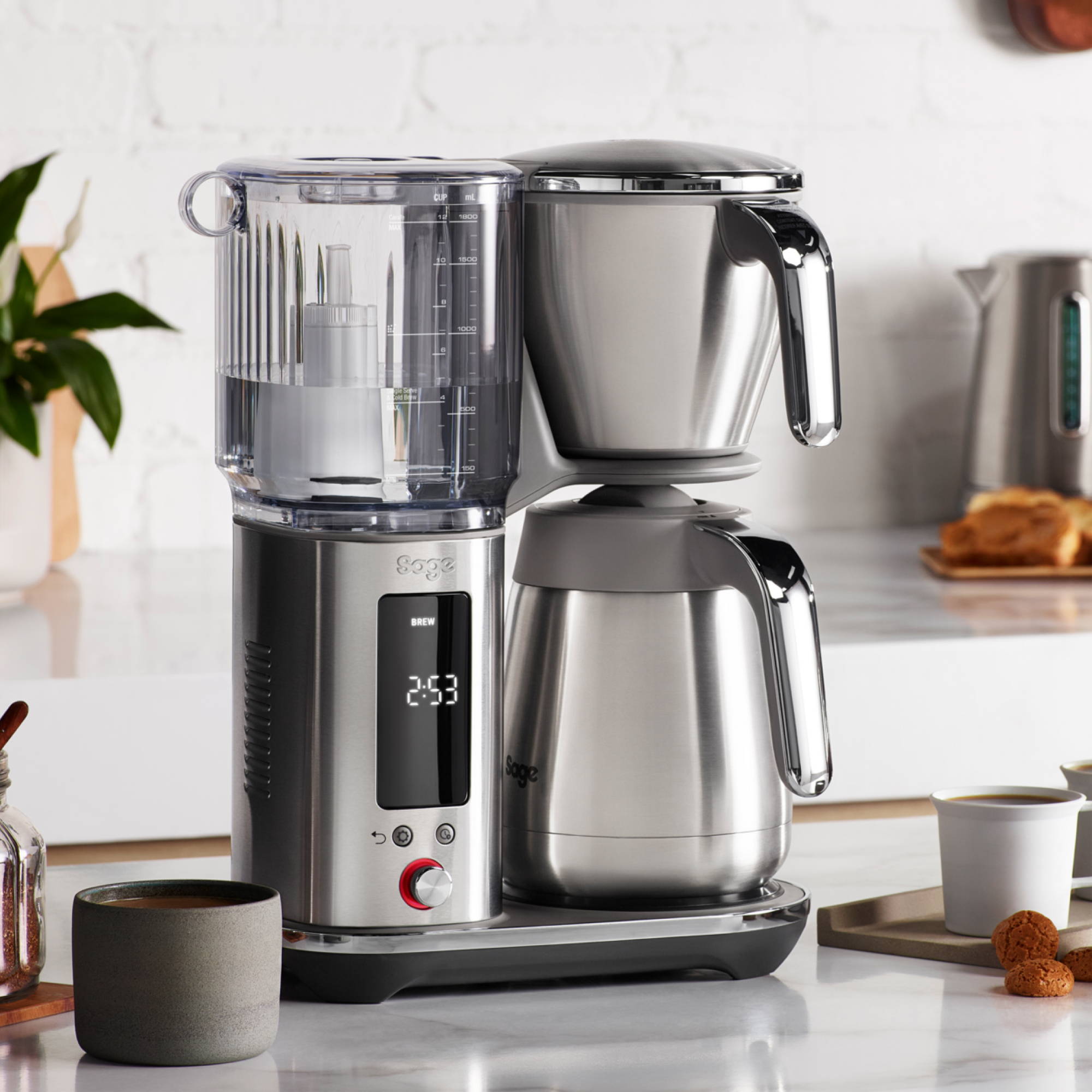 Eine vollständige Aufnahme der Sage Precision Brewer Kaffeemaschine aus gebürstetem Edelstahl, die auf einer Küchenarbeitsplatte steht. Man sieht den transparenten Wassertank auf der linken Seite, den Brühkorb auf der rechten Seite und eine Thermoskanne darunter. Ein digitales Display zeigt die Uhrzeit an. Im Hintergrund sind Küchenelemente wie eine Pflanze, Becher und ein Wasserkocher zu sehen.