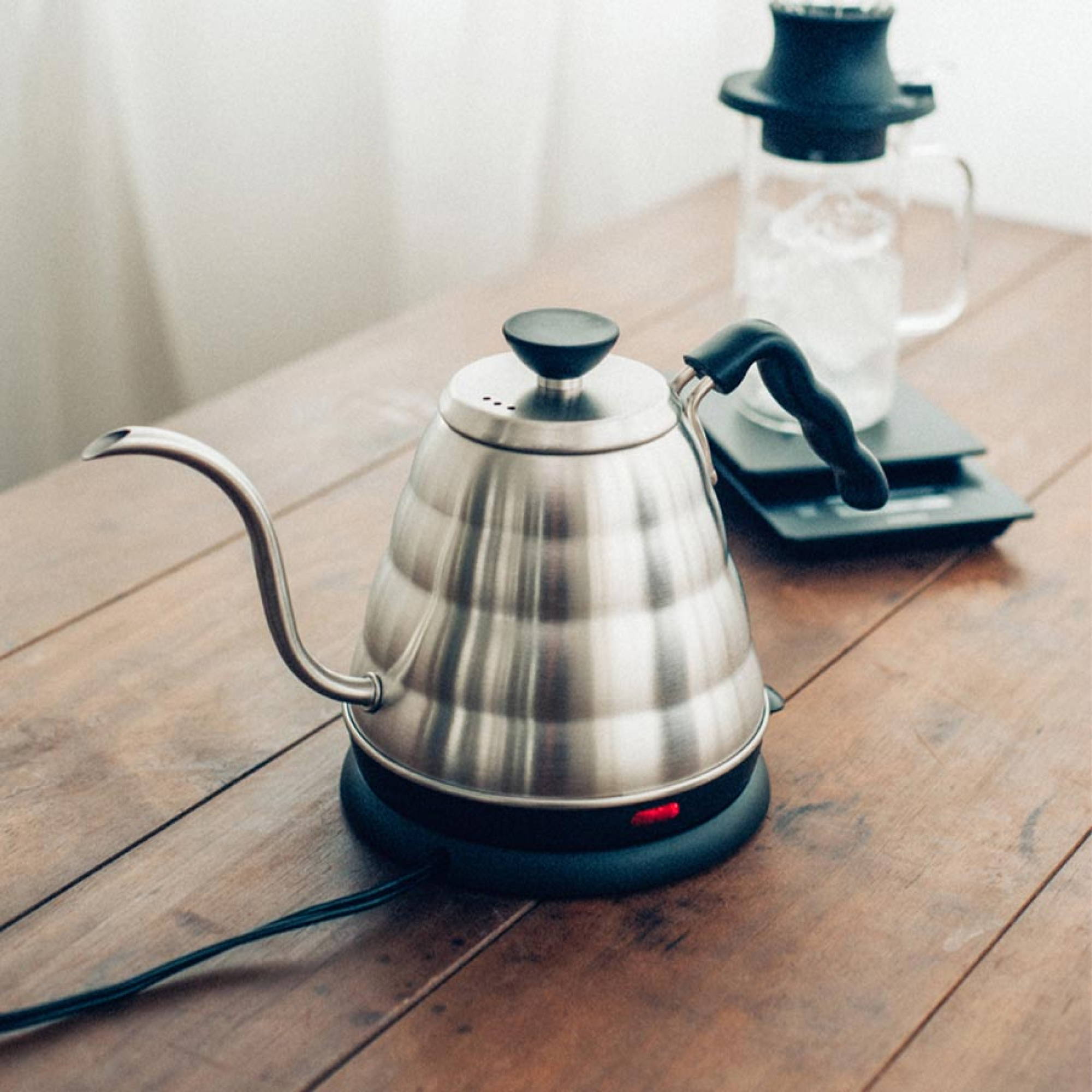 Elektrischer Hario V60 Buono Wasserkocher aus Edelstahl mit charakteristischem Schwanenhals-Ausguss auf einem Holztisch. Die rote Kontrollleuchte an der schwarzen Basis leuchtet. Im unscharfen Hintergrund stehen eine digitale Kaffeewaage und ein Handfilter-Set mit Glaskaraffe.