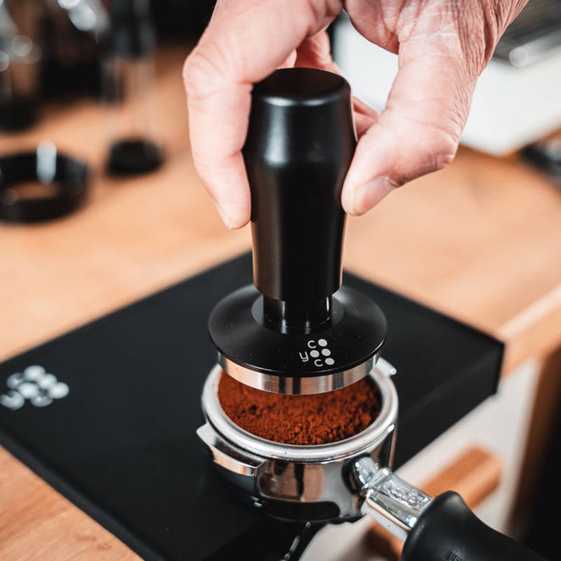 One hand holds a black Coyooco tamper to tamp coffee grounds in the portafilter.