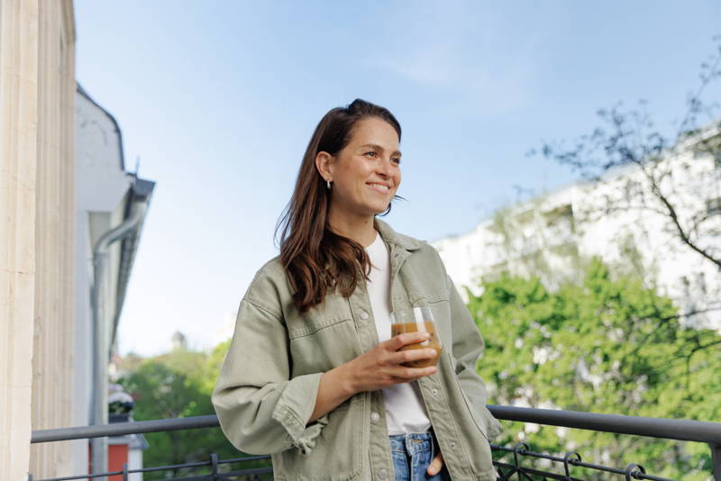 Frau steht auf Balkon und hält Iced Coffee in der Hand