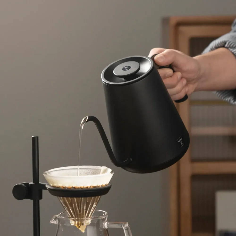 Eine Hand gießt heißes Wasser aus einem schwarzen Schwanenhals-Wasserkocher in einen Kaffeefilter auf einem Halter.