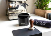 The Coffee Circle Barista Essential Set, consisting of a tamper, knock box, tamping mat, and cleaning cloth, is displayed on a light-colored table.