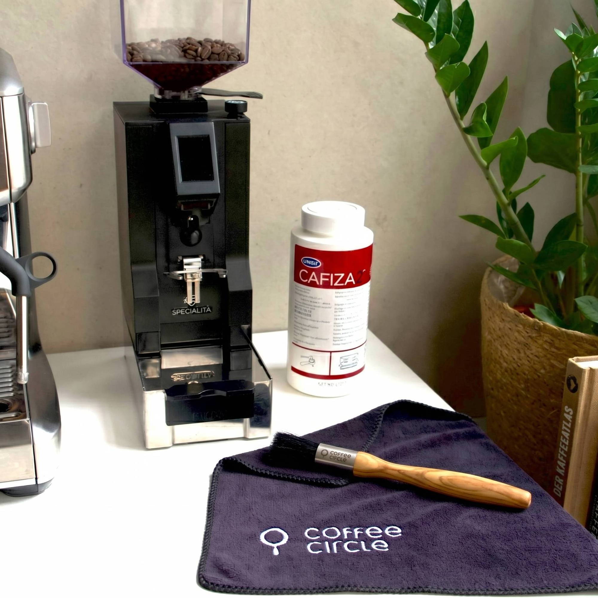  Cleaning kit for espresso machines on a white table next to a coffee grinder with coffee beans. On the table are a Coffee Circle microfiber cloth with a wooden brush and a can of Cafiza cleaner. In the background are a green houseplant and two coffee books, including “Good Coffee” by James Hoffmann.