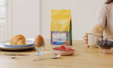 Auf einem Frühstückstisch steht eine Packung Warka-Kaffee, daneben liegen ein Croissant, ein Ei und Beeren.