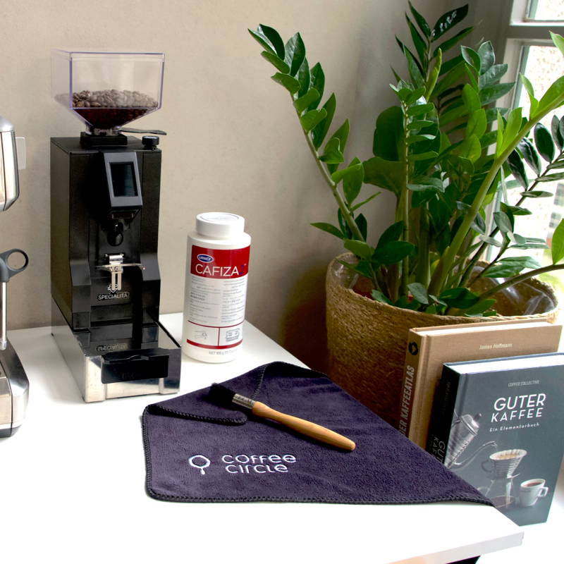 Cleaning kit for espresso machines on a white table next to a coffee grinder with coffee beans. On the table are a Coffee Circle microfiber cloth with a wooden brush and a can of Cafiza cleaner. In the background are a green houseplant and two coffee books, including “Good Coffee” by James Hoffmann.