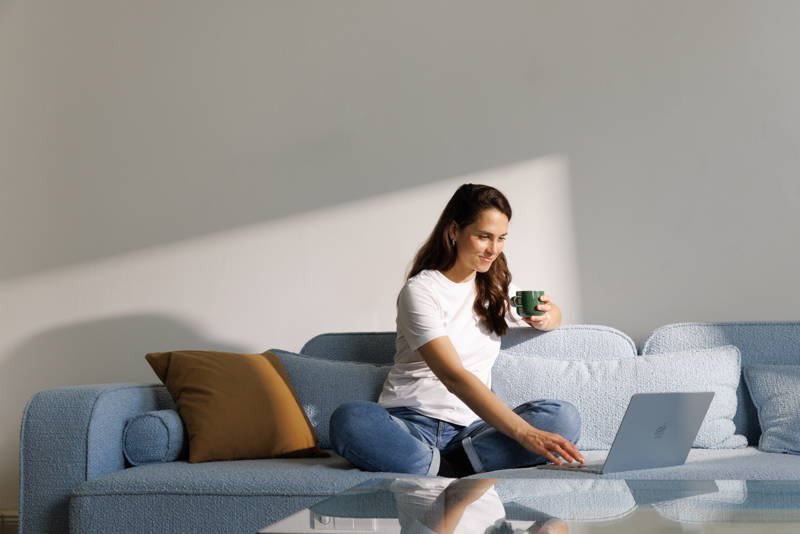 Woman sitting on a sofa, holding a cup of coffee while typing on a laptop