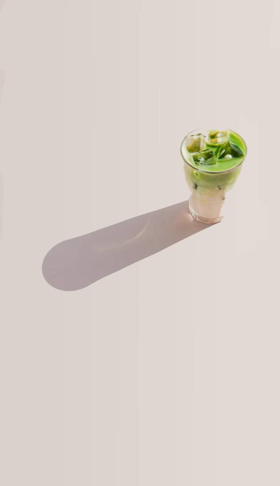 Minimalist product photography of an iced matcha latte in a glass. The drink features a distinct layer of milk at the bottom and vibrant green matcha on top, filled with ice cubes. Set against a clean, solid beige backdrop, the glass casts a long, smooth shadow.