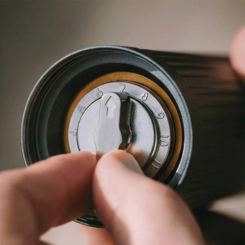 Eine Nahaufnahme der Unterseite der AeroPress Handkaffeemühle. Zwei Finger stellen den Mahlgrad an einem metallischen Drehregler mit nummerierter Skala ein (der Pfeil zeigt auf die 7).