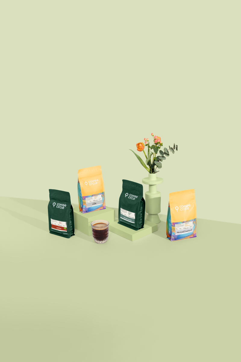 Das Frühlingskaffee Set mit unseren Espressos ist vor grünem Hintergrund aufgestellt. Im Vordergrund befindet sich ein Glas mit Kaffee und im Hintergrund eine Blumenvase mit orangenen Blumen