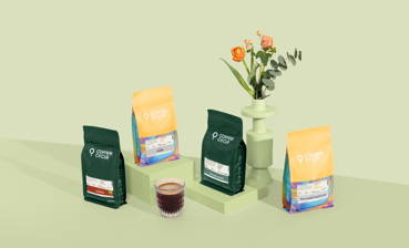 Das Frühlingskaffee Set mit unseren Espressos ist vor grünem Hintergrund aufgestellt. Im Vordergrund befindet sich ein Glas mit Kaffee und im Hintergrund eine Blumenvase mit orangenen Blumen